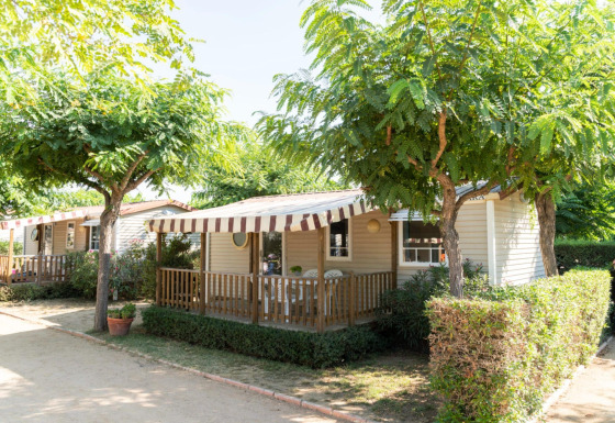 Casa móvil en Camping del Mar con porche y toldo a rayas, rodeada de árboles y arbustos verdes.
