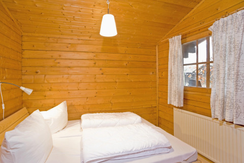 Slaapkamer in houten lodge met tweepersoonsbed, witte lakens en raam in Type C House, Duitsland.