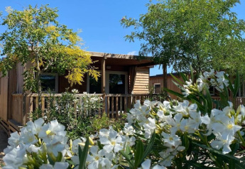 Casa móvil con terraza de madera rodeada de árboles verdes y flores blancas en Els Pins Mobile-Home Plus 2.