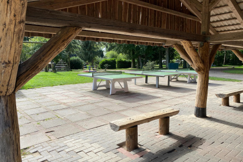 Outdoor ping pong tables under a wooden shelter with benches, surrounded by green lawns and playground in Germany.