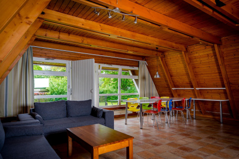 Lodge interior with wooden beams, a gray sofa, wooden table, and colorful chairs by large windows.