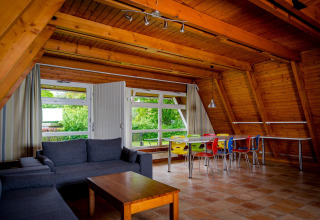 Warme leefruimte in een houten lodge met grijze zetel, houten tafel en kleurrijke stoelen aan het raam.