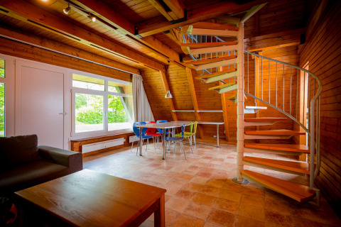 Bright lodge interior with wooden walls, spiral staircase, colorful dining chairs, and a large window view.