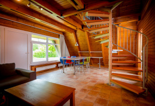 Lichte lodge met houten wanden, wenteltrap, kleurrijke stoelen rond tafel, en groot raam met tuinzicht.
