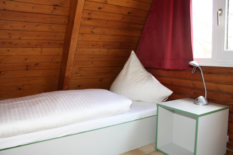 Een eenpersoonsbed bij het raam in een houten lodge met een rood gordijn en een witte lamp.