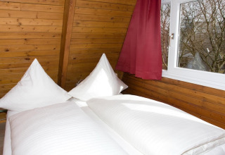 Slaapkamer in Nurdachhaus Saver Rate 65 m² in Duitsland, houten muren, wit beddengoed en raam met gordijn.