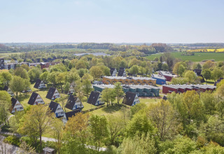 Luchtfoto van een lodge met A-frame huisjes en kleurrijke blokhuizen tussen groene bomen en velden.