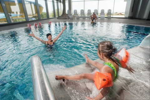 Indendørs swimmingpool med glade børn, hvor en pige med orange badevinger leger ved kanten på en lodge.