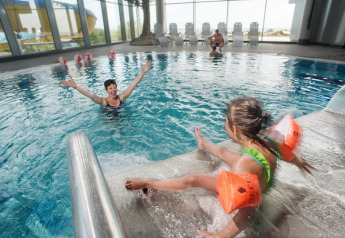 Piscina cubierta en un lodge con una niña usando flotadores naranjas jugando en los escalones cerca de un adulto.