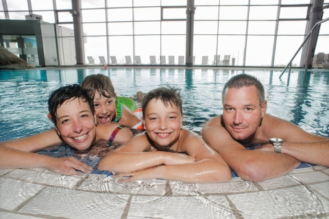 En glad familie på fire slapper af sammen i en indendørs pool på et moderne lodge med store vinduer.