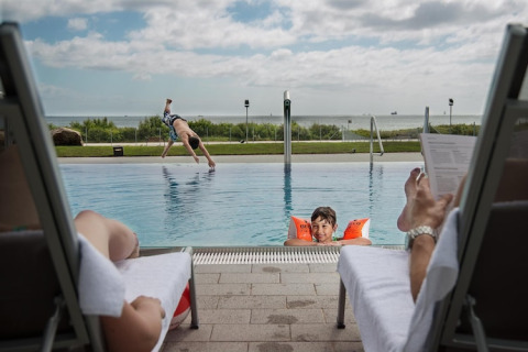 Twee volwassenen relaxen bij het zwembad, een kind zwemt met zwembandjes, een ander springt erin.