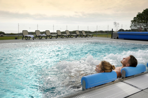 Two people relaxing in a bubbling outdoor lodge pool, with lounge chairs lined up in the background at sunset.