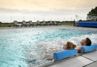 Two people relaxing in a bubbling outdoor lodge pool, with lounge chairs lined up in the background at sunset.