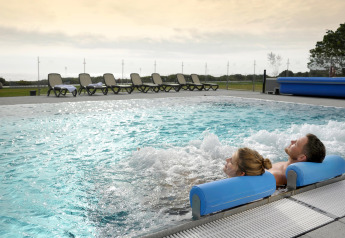 Dos personas se relajan en una piscina exterior con burbujas en un lodge, con tumbonas al fondo al atardecer.