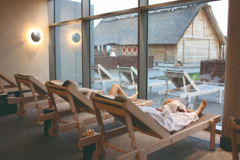 Mensen ontspannen op ligstoelen in een lodge met grote ramen en uitzicht op houten huisjes buiten.