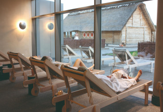 Mensen rusten op ligzetels binnen in een lodge met grote ramen en uitzicht op houten hutten buiten.