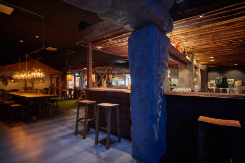 Rustiek lodge-interieur met houten balken, stenen zuilen, barzitjes en warme sfeerverlichting in de avond.