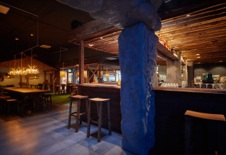 Rustiek lodge-interieur met houten balken, stenen zuilen, barzitjes en warme sfeerverlichting in de avond.