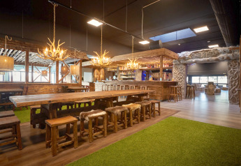 Cozy lodge interior with long wooden tables, antler chandeliers, and rustic decor by the bar area.