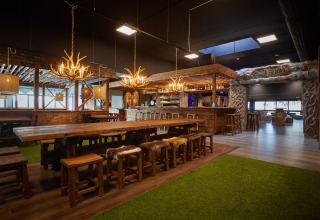 Cozy lodge interior with long wooden tables, antler chandeliers, and rustic decor by the bar area.