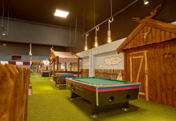 Albergue interior con alfombra verde, mesa de billar, futbolín y cabañas rústicas de madera decoradas.