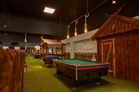Indoor lodge met groen tapijt, pooltafel, tafelvoetbal en rustieke houten hutten in gezellige sfeer.