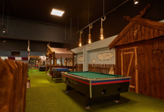 Indoor lodge met groen tapijt, pooltafel, tafelvoetbal en rustieke houten hutten in gezellige sfeer.