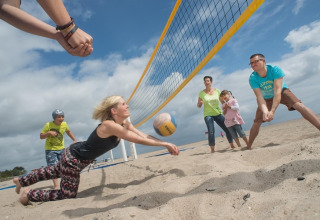 Familie spiller beachvolley ved et lodge under blå himmel, alle smiler og nyder en aktiv dag sammen.