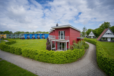 Rødt terrassehus i Ostseeresort Dampland, Tyskland, omgivet af grønne hække og farverige huse.