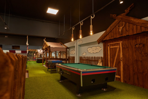 Sfeervolle lodge-ruimte met biljarttafel, groene vloerbedekking, houten hutten en warme verlichting.