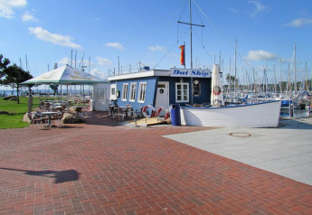 Outdoor café by a marina with many sailboats, blue building with 'Dat Ship' sign, sunny day.