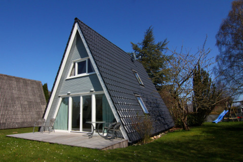 A-formet feriehus i Tyskland med terrasse og havemøbler på en solrig dag med blå himmel.