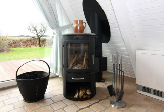 Moderne houtkachel met gereedschap en houtmand naast groot raam in vakantielodge Damp, Duitsland.