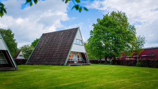 Een foto van een modern A-vormig huisje op een groen gazon, omringd door bomen en deels bewolkte lucht.