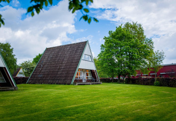 A photo of a modern A-frame lodge on a green lawn, surrounded by trees and under a partly cloudy sky.