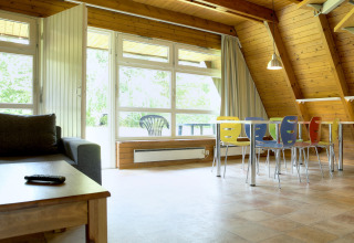 Lichte, ruime lodge woonkamer met kleurrijke stoelen rond een tafel, grote ramen en houten wanden.