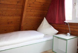 Eenpersoonskamer in houten lodge met bed, rood gordijn en raam in Nurdachhaus, Duitsland.