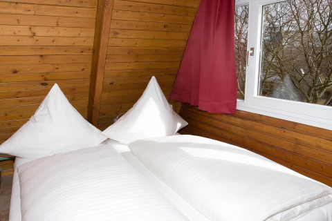 Een knus bed met witte kussens en dekbed naast het raam in een houten kamer met rode gordijnen.