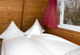 Een gezellig bed met witte kussens en dekbed naast het raam in een kamer met houten wanden en rode gordijnen.