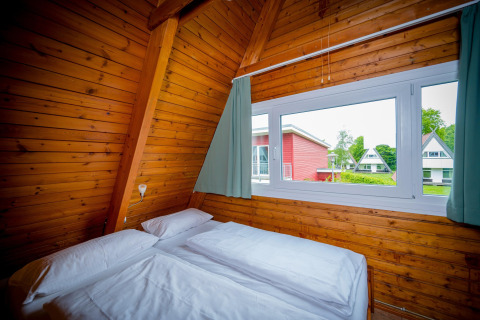 Slaapkamer in houten lodge met tweepersoonsbed en groot raam met uitzicht op andere lodges buiten.