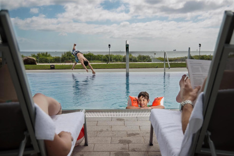 Twee volwassenen ontspannen bij het zwembad van een lodge, terwijl kinderen spelen en duiken in het water.