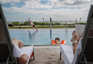 Twee volwassenen ontspannen bij het zwembad van een lodge, terwijl kinderen spelen en duiken in het water.