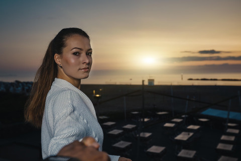 Woman in a white sweater stands at a lodge during sunset, with beach seating and calm sea behind her.