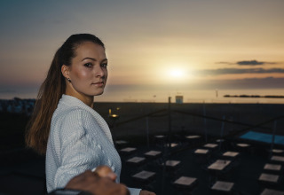 Woman in a white sweater stands at a lodge during sunset, with beach seating and calm sea behind her.