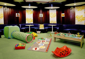 Salle de jeux pour enfants dans un lodge avec tunnel, tapis coloré, blocs et banquettes autour de la pièce.