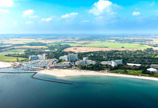 Luchtfoto van een kustgebied met lodge, strand, jachthaven en groene velden op een zonnige dag.