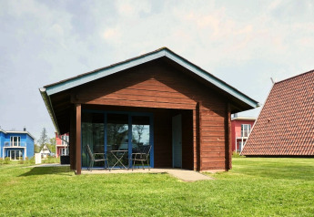Cabaña de madera con terraza cubierta y muebles exteriores, rodeada de césped y casas vecinas de colores.