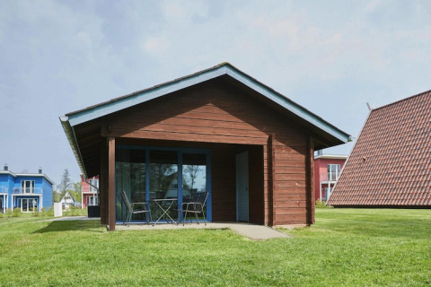 Lodge di legno con patio coperto e mobili da esterno, circondato da prato e case colorate nelle vicinanze.