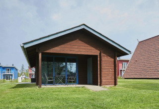 Træhytte med overdækket terrasse og havemøbler i grønne omgivelser tæt på andre farverige huse.