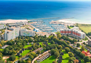 Vue aérienne d’un port de plaisance avec bateaux, hôtels et chalets près de la mer sous ciel bleu.